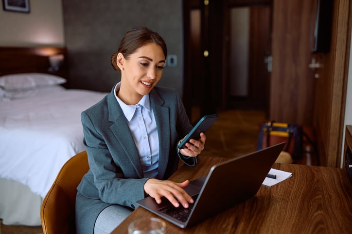 Business woman working on a laptop while holding a cellular phone in a hotel room. Hotel room design for bleisure.